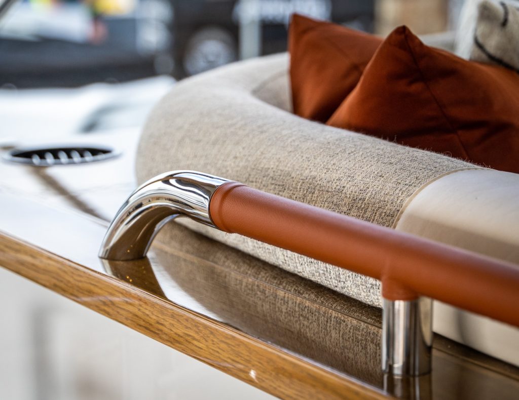 Up close of chrome railing with a burnt orange leather wrapping, next to a grey fabric sofa adorned with burnt orange scatter cushions, all inside a yacht